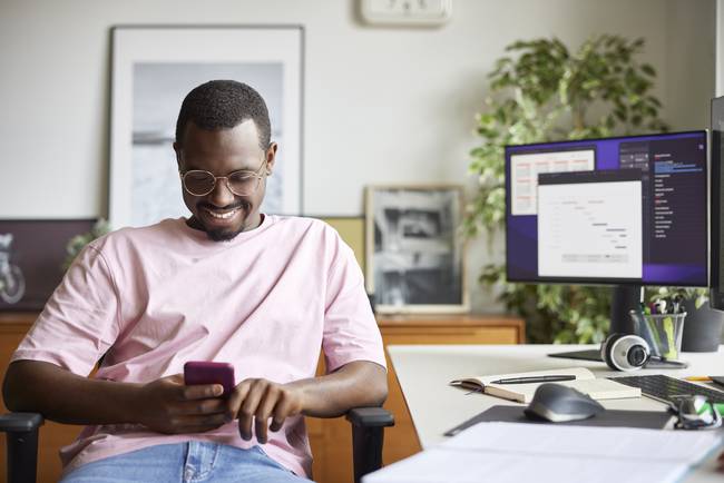 Hombre sonriendo a su celular con un computador de escritorio de fondo (Getty Images)